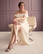 Woman in a white off-shoulder dress sitting on a chair against a purple paneled wall. This dress is the Ellington Strapless Dress sewing pattern from Cashmerette.