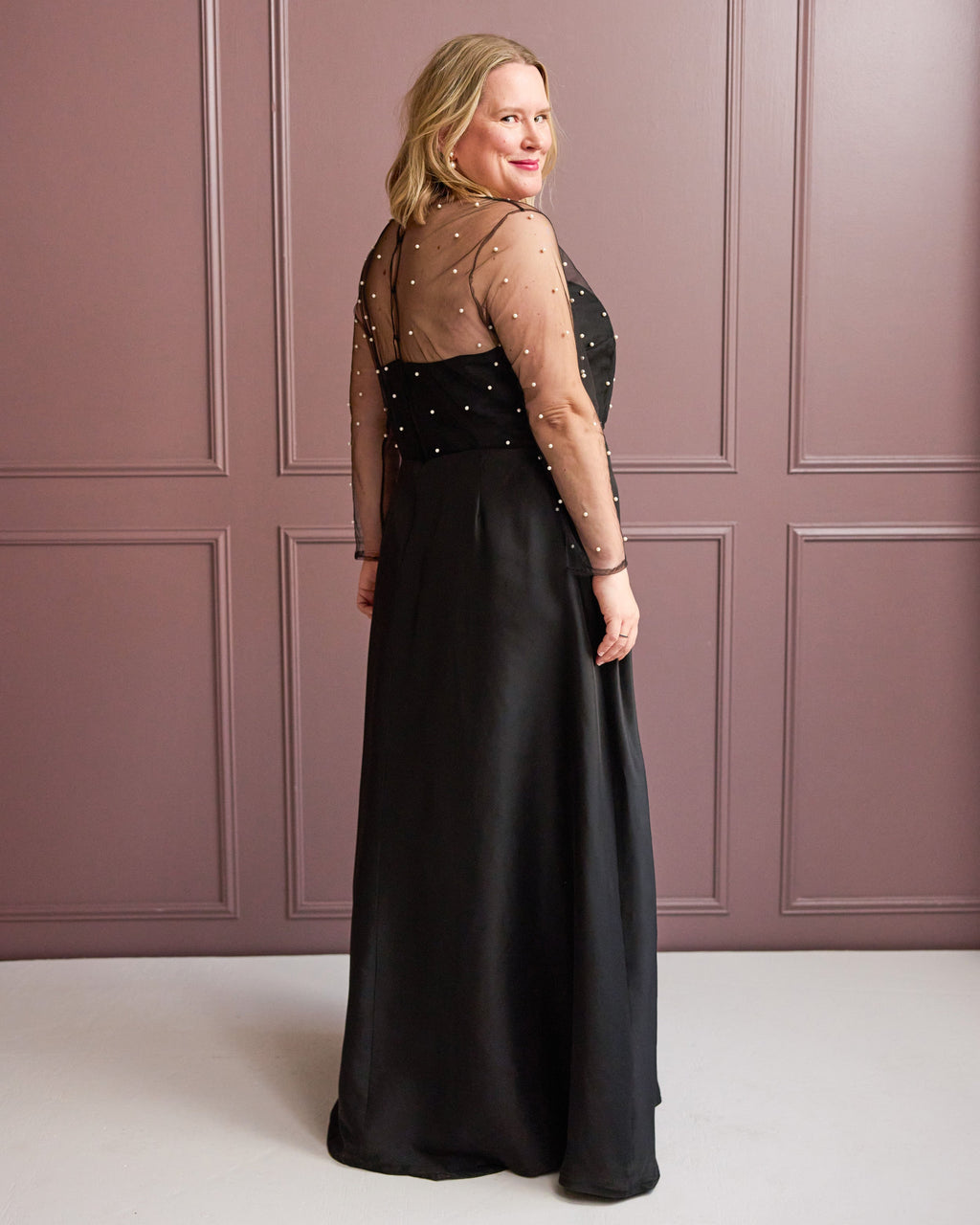 Woman wearing a black evening gown with sheer sleeves against a pink paneled wall. This is the Ellington Dress sewing pattern from Cashmerette.