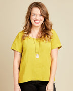 Woman wearing a yellow shirt and black pants against a beige background. This top is the Montrose Top sewing pattern from Cashmerette.