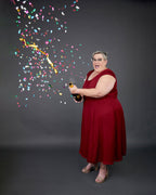 Woman in a red dress holding a bottle with confetti falling around her on a dark background. This dress is the Upton Dress & Skirt sewing pattern from Cashmerette.