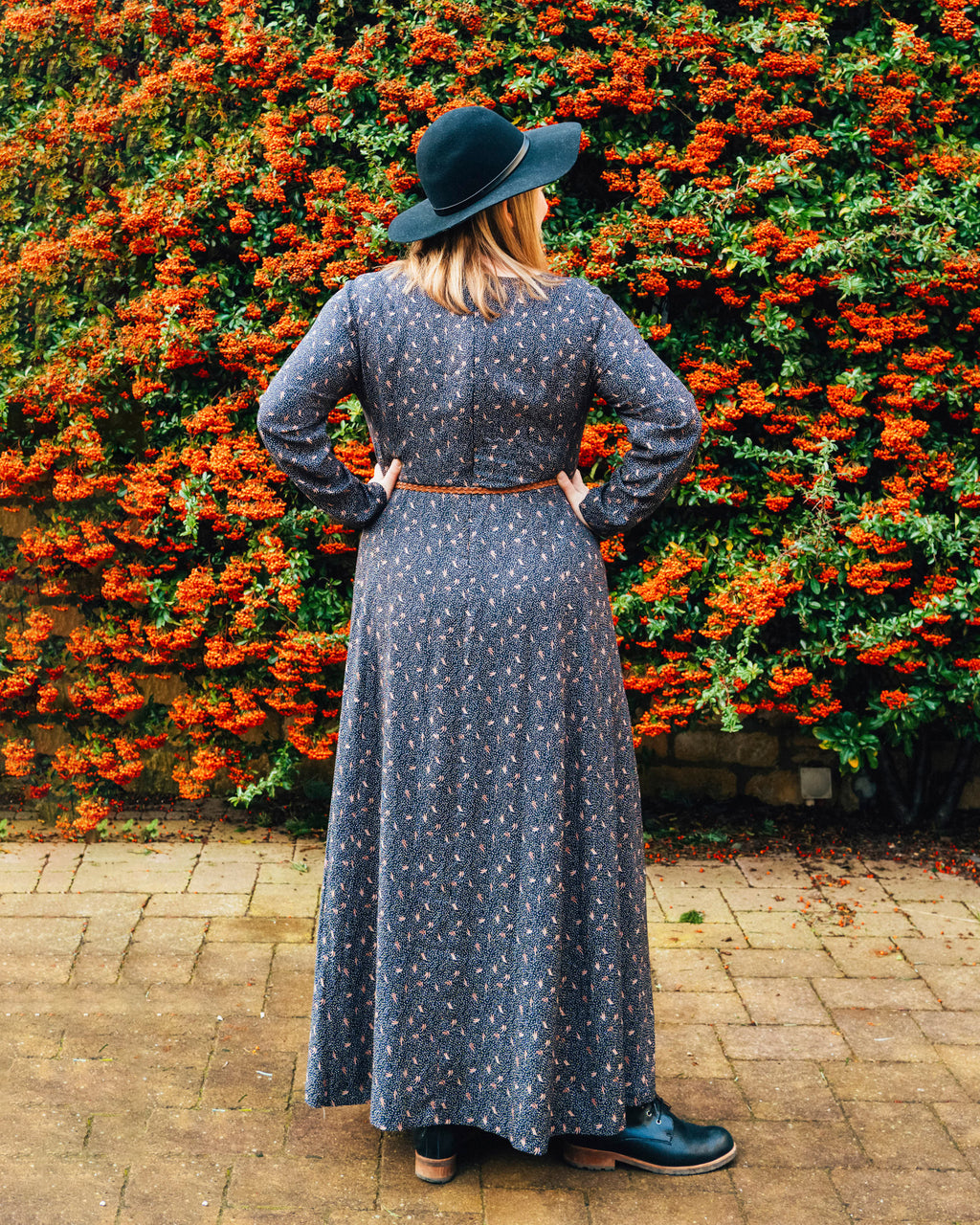 Woman in a long dark dress and black hat standing in front of a wall of orange flowers.. This dress is the Upton Dress & Skirt sewing pattern from Cashmerette.