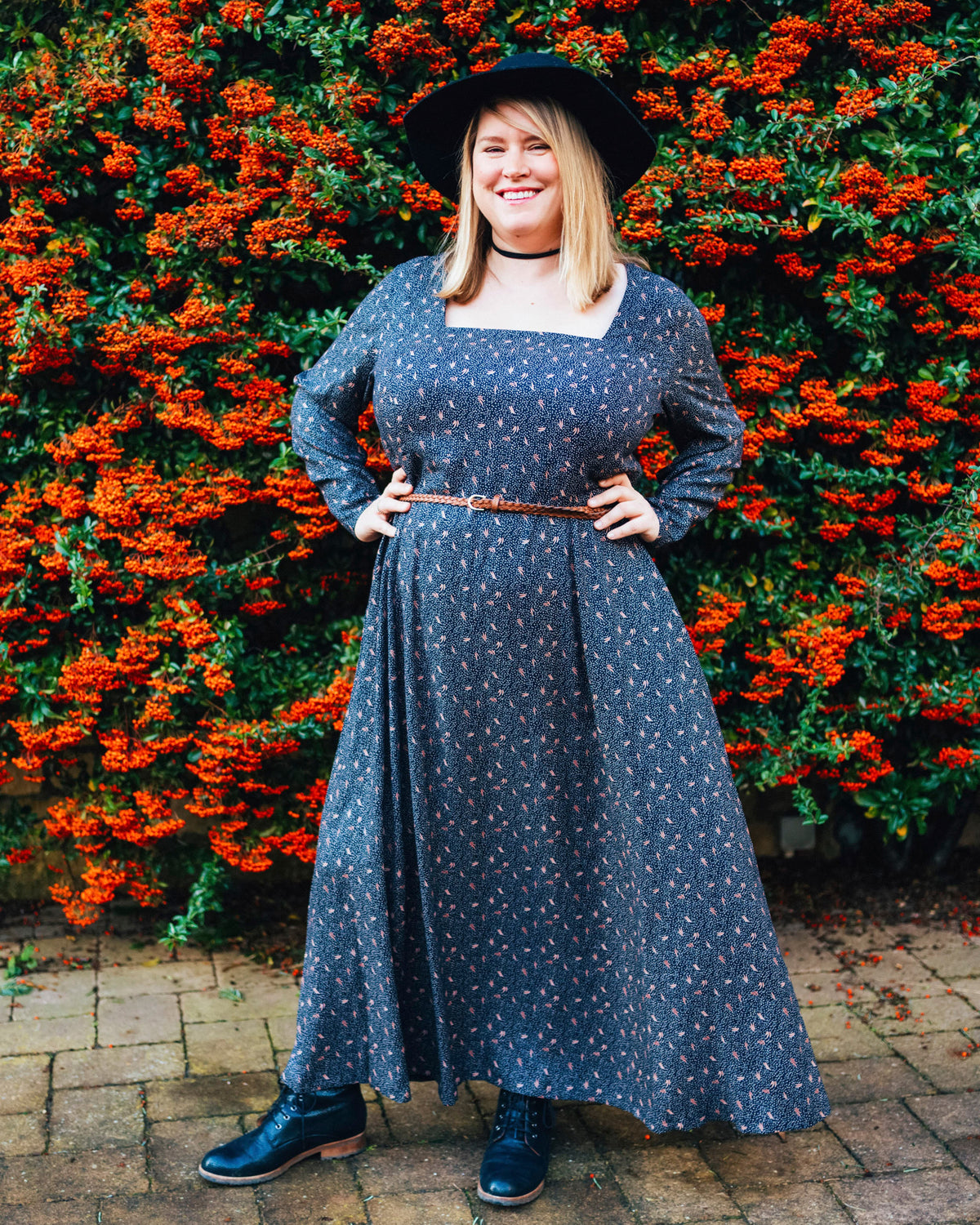 Woman in a blue dress standing in front of a hedge with red flowers. This dress is the Upton Dress & Skirt sewing pattern from Cashmerette.